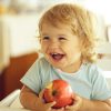 Laughing cute fair-haired blond hazel-eyed kid little child baby boy sitting in highchair and eating big red apple fruit portrait on blurred background horizontal picture