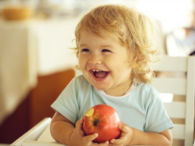 Laughing cute fair-haired blond hazel-eyed kid little child baby boy sitting in highchair and eating big red apple fruit portrait on blurred background horizontal picture