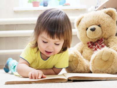 little girl reading book lying on stomach in her room on carpet with toy Teddy bear smiling cute child children education and development happy childhood